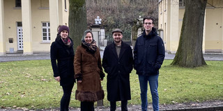 Zwei Frauen und zwei Männer in Winterjacken stehen nebeneinander und schauen in Richtung Kamera. Im Hintergrund Wiese, zwei Bäume und Gebäude.