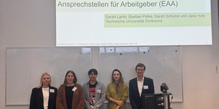 Drei Frauen und zwei Männer stehen hinter einem Hörsaalpult und schauen in Richtung Kamera.